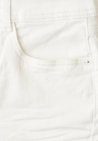 Gros plan sur un tissu en denim blanc montrant le coin supérieur d'une poche avant avec des coutures et un petit rivet métallique.