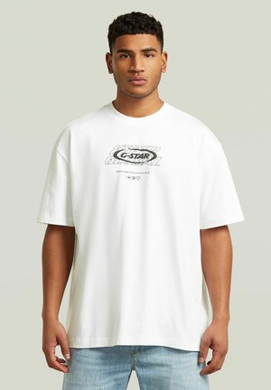 Mężczyzna ubrany w luźny biały t-shirt z czarnym napisem i jasnoniebieskie dżinsy, stojący na tle jednolitego jasnego tła.