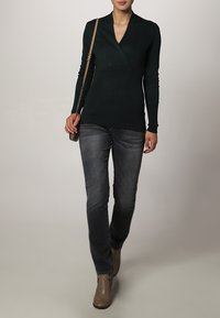 Mørkegrøn omslagstrøje i blødt strik, med en dyb v-hals og lange ærmer, parret med tætsiddende grå jeans og beige ankelstøvler.