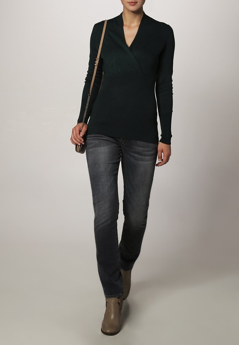Mørkegrøn omslagstrøje i blødt strik, med en dyb v-hals og lange ærmer, parret med tætsiddende grå jeans og beige ankelstøvler.