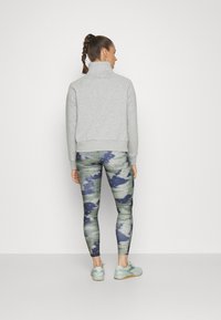 Svijetlosivi dukserica od flisa s visokim ovratnikom, uparen s leginsima sa uzorkom u zelenim, plavim i bijelim nijansama. Svijetle tenisice upotpunjuju outfit.