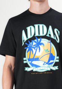 Fekete póló színes grafikai tervezéssel, rajta pálmafákkal, egy vitorlással és a nappal, feliratozva: "ADIDAS ORIGINALS VACATION SEASON."