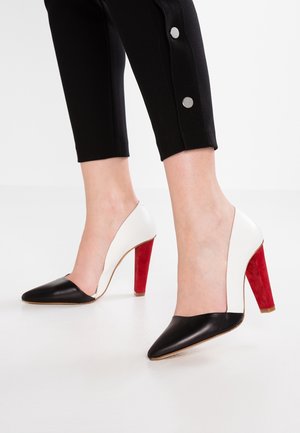Czarno-białe buty na wysokim obcasie z czerwonym zamszowym obcasem. Projekt charakteryzuje się szpiczastym noskiem i wycięciem w kształcie litery V po bokach.
