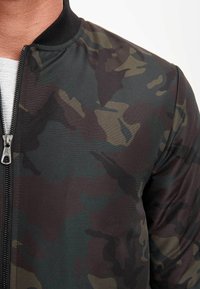 Camo bomber jacket with a black ribbed collar, featuring dark green, brown, and gray camouflage patterns, and a silver zipper closure.