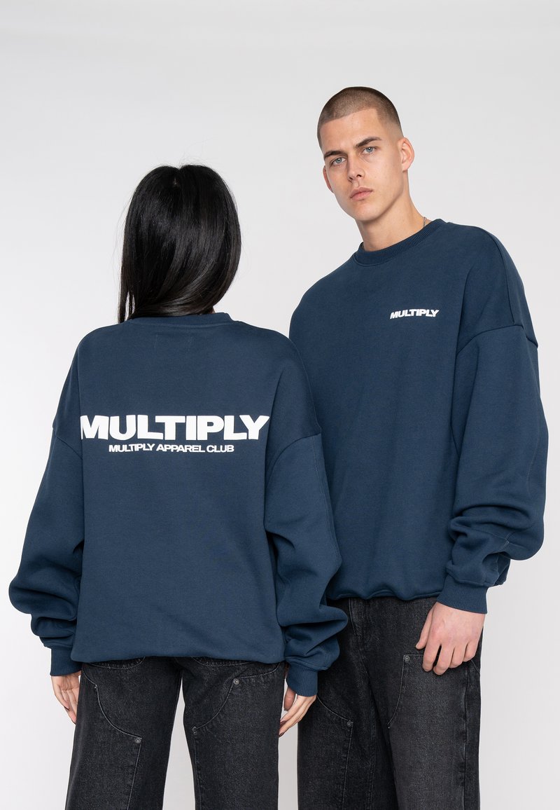 Felpa oversize blu navy con testo bianco "MULTIPLY" e "MULTIPLY APPAREL CLUB" sul retro. Maniche lunghe e polsini a costine.
