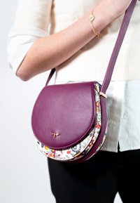 Borsa a tracolla viola in pelle con tessuto a motivo floreale sul lato inferiore. Presenta un design curvo e un dettaglio in accento dorato.