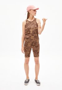 Bruine tie-dye tanktop en korte broek set, met een aansluitend ontwerp. Het model draagt een roze pet en grijze sneakers, waarbij ze poseert met één hand omhoog.