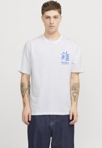 Camiseta de algodón blanca con gráficos impresos en azul en el pecho. Mangas cortas y un corte relajado, combinada con pantalones de mezclilla oscuros.
