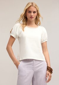 Top alb cu mâneci scurte, volanitate, gât rotund și tiv contrastant. Modelul este asortat cu pantaloni violet deschis și brățări din lemn.