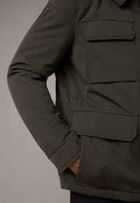 Olivenfarbene Jacke mit glatter Textur, ausgestattet mit zwei Brusttaschen und einer Seitentasche, strukturierten Schultern und einem Reißverschluss.