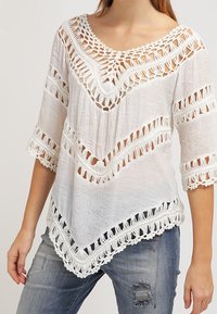 Haut en crochet blanc avec décolleté en V et manches trois-quarts, associé à un jean bleu déchiré, porté par une personne debout.