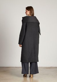 Abrigo largo de lana gris oscuro con cuello ancho, detalle de lazo en la espalda y corte ajustado, combinado con pantalones oscuros holgados y zapatos de tacón alto.