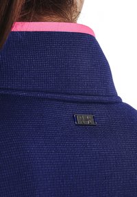 Námořnická modrá texturovaná tkanina s vysokým límcem, se světle růžovým lemem a malou černou štítkovou nálepkou s emblémem na zadní straně.