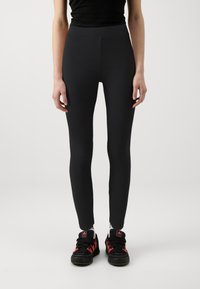 Leggings noirs en tissu lisse et extensible avec une taille haute, associés à des chaussures de sport noires et rouges.