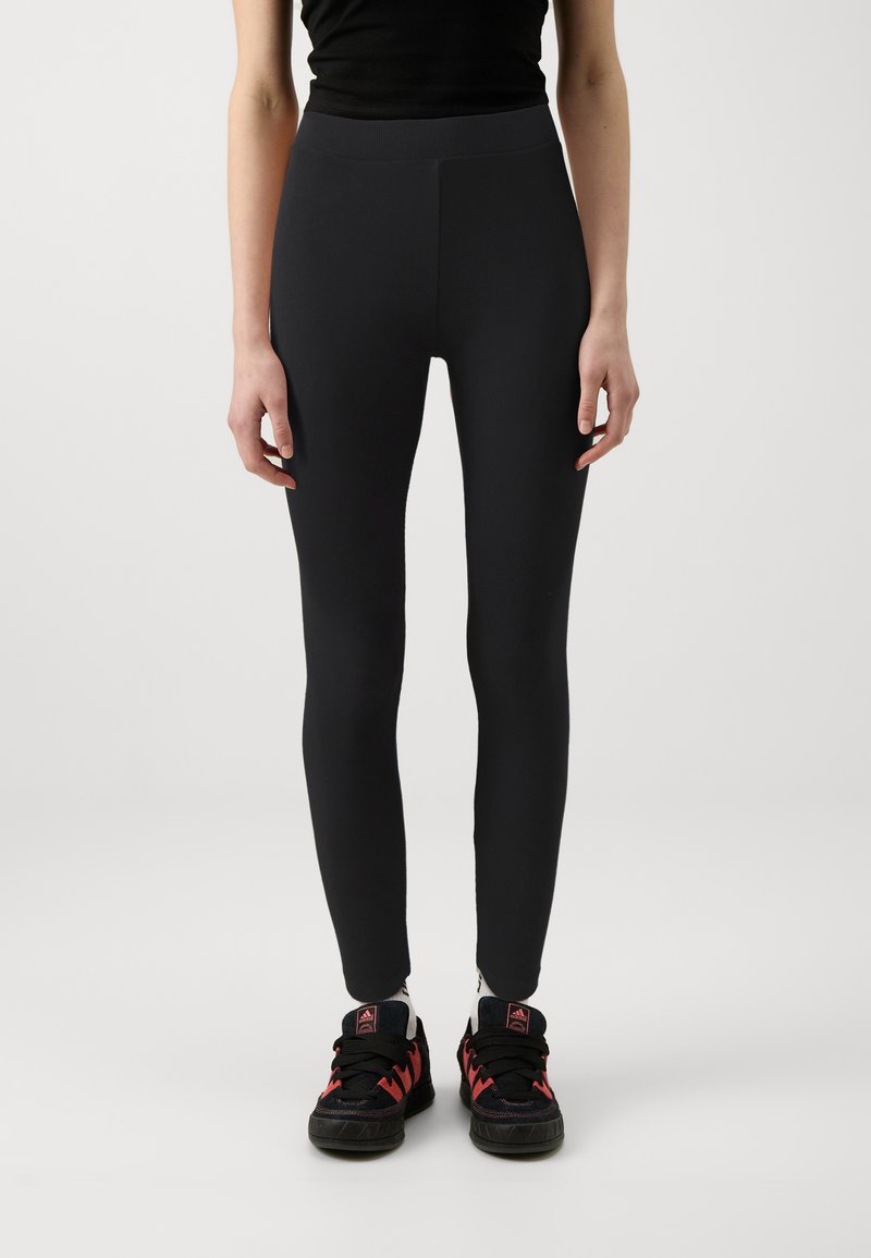 Leggings noirs en tissu lisse et extensible avec une taille haute, associés à des chaussures de sport noires et rouges.
