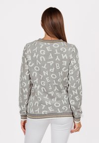 Donna con lunghi capelli castani che indossa un maglione grigio con lettere bianche sparse e pantaloni bianchi, rivolta di spalle alla fotocamera.