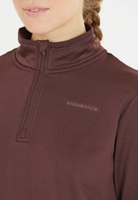 Burgunderfarbenes Langarmoberteil mit hohem Kragen, das über einen halben Reißverschluss, strukturiertes Material und ein kleines "ENDURANCE"-Logo auf der Brust verfügt.