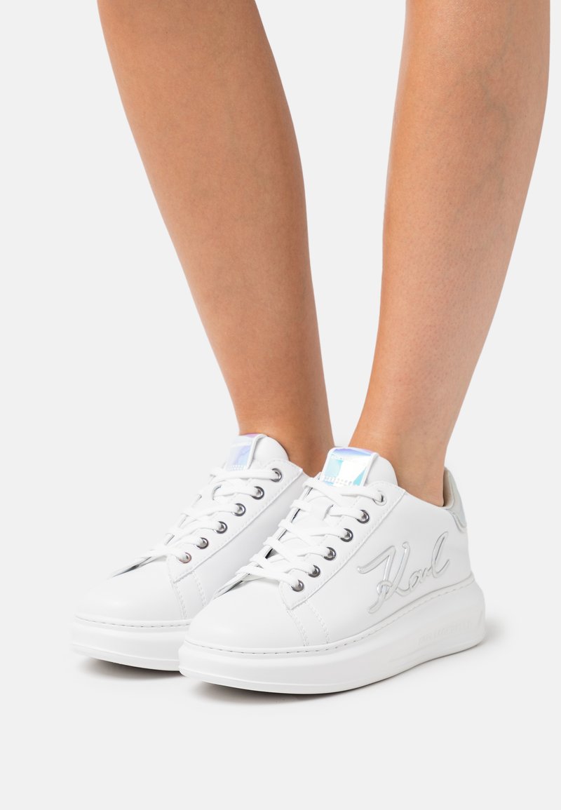 Baskets en cuir blanc avec une texture lisse, présentant des accents de logo argentés, un bout arrondi et une semelle en caoutchouc blanche. Les lacets sont des œillets en métal standard.