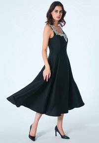 Robe noire sans manches avec une jupe évasée et des détails de col en perles, fabriquée en tissu lisse, assortie à des chaussures à talons hauts noires.