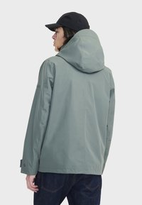Veste imperméable vert clair avec capuche, présentant une coupe décontractée, une texture lisse et des poignets à boutons-pression. La vue arrière montre un design minimaliste.