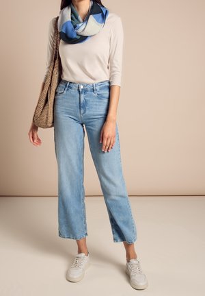 Femme portant un jean large bleu clair, un haut beige à manches longues, une écharpe bleue à motifs, des baskets blanches et portant un sac tressé marron.