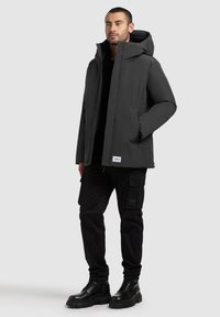 Black insulated jacket with a hood, featuring a front zipper and side pockets. Paired with black cargo pants and heavy black lace-up boots.