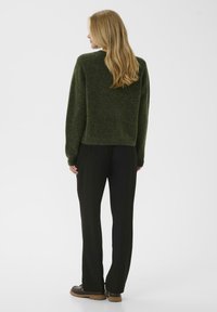 Zaļš vilnas džemperis ar brīvu piegriezumu un ribotu apakšmalu, kombinēts ar melniem biksēm un brūnām kurpēm, skatīts no aizmugures.