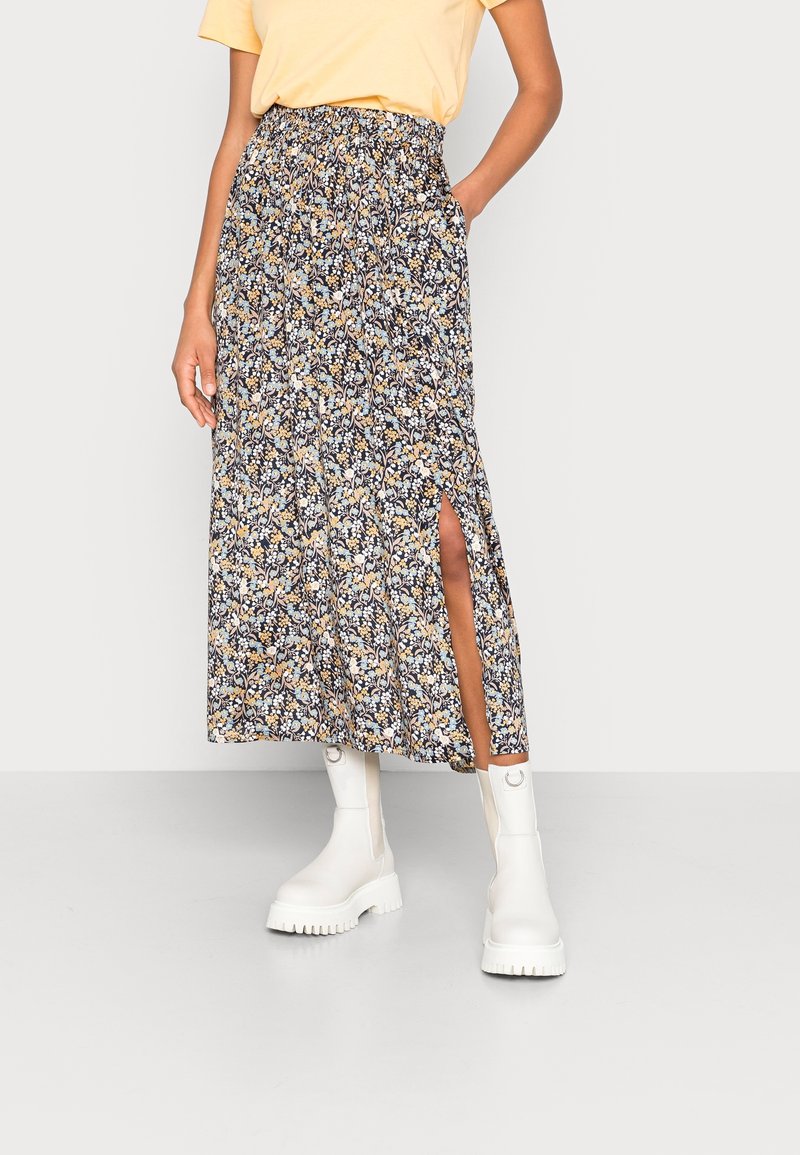 Femme portant une jupe midi fleurie avec une fente latérale, une chemise jaune et des bottes blanches épaisses, debout avec une main dans la poche.