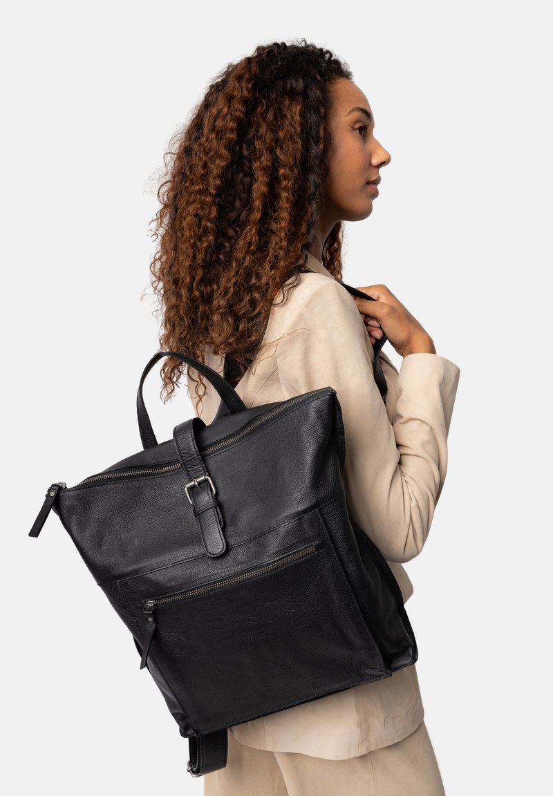 Zaino in pelle nera con finitura testurizzata, manico superiore, spallacci regolabili e tasca frontale con zip. Modello mostrato con una giacca chiara.