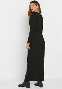 Robe maxi noire côtelée avec des manches longues et un détail de ceinture, présentant une silhouette ajustée et un col rond, associée à des bottines.