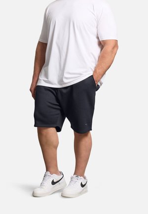 Mężczyzna w białej koszulce z krótkim rękawem, czarnych krótkich spodenkach do kolan i białych butach sportowych Nike stoi z rękami w kieszeniach.