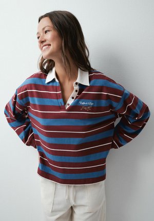 Femme souriante aux cheveux foncés portant un maillot de rugby bleu et bordeaux rayé avec col blanc et pantalon blanc, debout devant un mur uni.