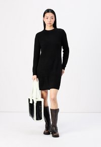 Vestido negro de punto con mangas largas, de largo hasta el muslo. La modelo sostiene un bolso de piel sintética en blanco y negro. Botas marrones hasta la rodilla completan el conjunto.