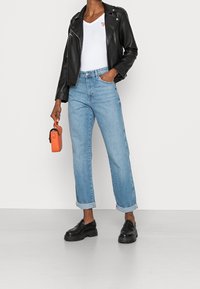 Czarna skórzana kurtka motocyklowa, biała koszulka z dekoltem w serek, jasnoniebieskie spodnie z wywiniętymi nogawkami, czarne masywne loafersy i pomarańczowa torba.