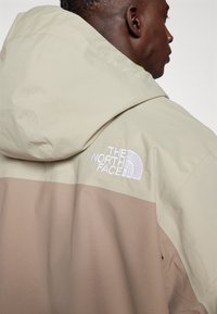 Beige og brun to-farget jakke med hette, laget av teksturert stoff, med "The North Face"-logo brodert i hvitt på skulderen.
