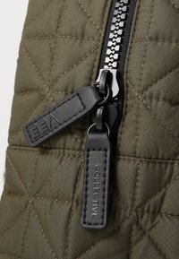 Tissu matelassé vert olive avec une finition texturée, comportant une fermeture éclair noire et des tirettes en cuir embossées avec "EEA" et "COLLECTIVE."