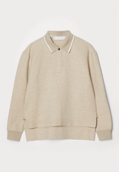Sweatshirt polo bege com colarinho e fecho de botão. Textura macia, fendas laterais e contornos brancos em contraste à volta do colarinho.