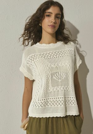 Jauna moteris su garbanotais plaukais, apsirengusi balta trumpomis rankovėmis megzta palaidine su raštuotu dizainu ir alyvuogių žalios spalvos kelnėmis, stovinti prie tekstūruotos sienos.