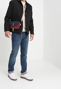 Svart jacka över en gräddfärgad tröja, blå jeans och vita sneakers. En röd och marinblå crossbody-väska med en rem hänger från axeln.