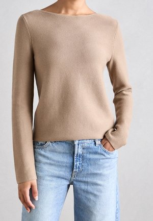 Kobieta ubrana w beżowy sweter z długim rękawem wykonany z dzianiny oraz jasnoniebieskie dżinsy, z jedną ręką w kieszeni, na neutralnym tle.