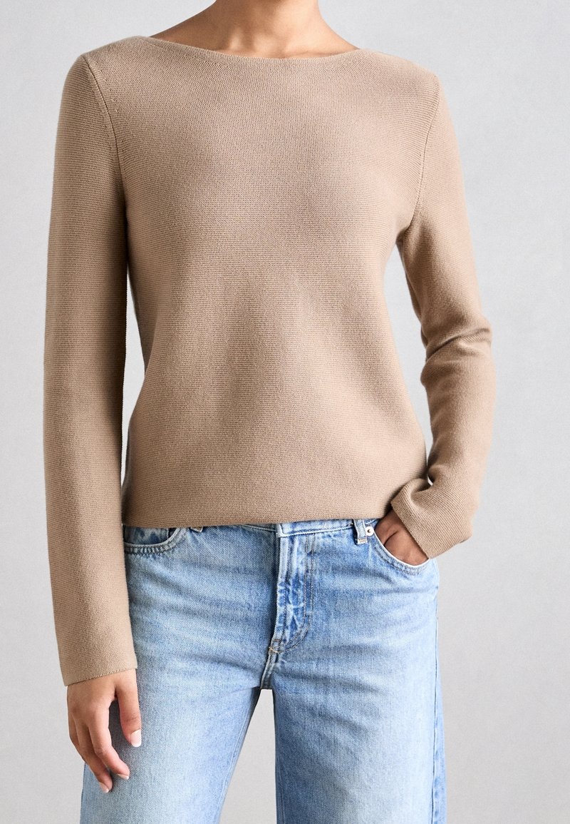 Femme portant un pull en maille à manches longues beige et un jean en denim bleu clair, avec une main dans la poche, sur un fond neutre.