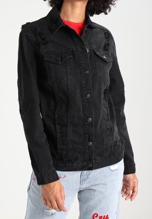 Henkilö, joka on pukeutunut mustaan nappilistalliseen farkkutakkiin punaisen paidan ja vaaleansinisten revittyjen farkkujen, joissa on punainen kirjailu, päällä, seisoo valkoista taustaa vasten.