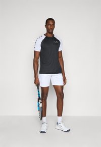 Svart og hvit sportsskjorte med korte ermer, sammen med hvite shorts. Modellen holder en tennisracket, ikledd hvite treningssko.