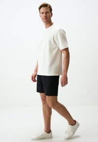 Chemise blanche à manches courtes avec un col rond, associée à un short noir. Le mannequin porte des baskets blanches. Texture lisse sur l'ensemble.