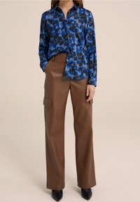 Blusa a fiori blu con maniche lunghe, abbinata a pantaloni cargo larghi in finta pelle marrone con tasche laterali e eleganti tacchi neri.