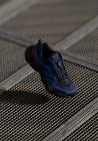 Der Merrell Matts Schuh zeichnet sich durch ein blau-schwarzes Design, ein strukturiertes Mesh-Obermaterial, schwarze Akzente und eine pinke Sohle aus. Er verfügt über GORE-TEX Branding.
