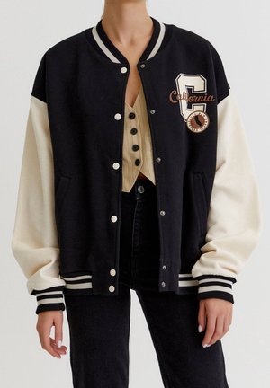 Veste varsity noire et crème avec écusson « California », portée sur un top beige à boutons et un pantalon noir, vue de face du cou aux hanches.