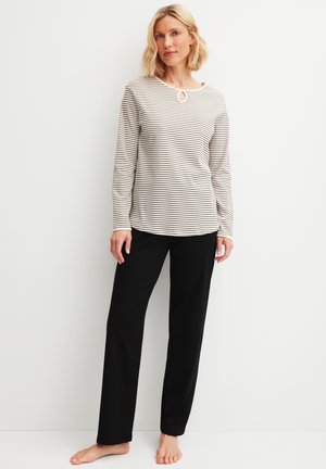 Donna in piedi a piedi nudi che indossa una maglietta bianca a maniche lunghe con sottili strisce nere e pantaloni neri a gamba dritta su sfondo bianco.