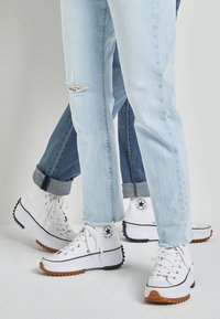 Białe sneakersy z wysoką cholewką i czarnymi akcentami, z teksturowaną gumową podeszwą, w komplecie z jasnymi i ciemnymi dżinsami, które mają postrzępiony dół.