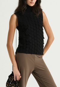 Haut noir sans manches avec un design texturé et superposé, doté d'un col montant. Associé à un pantalon ajusté de ton neutre et un sac décoratif.